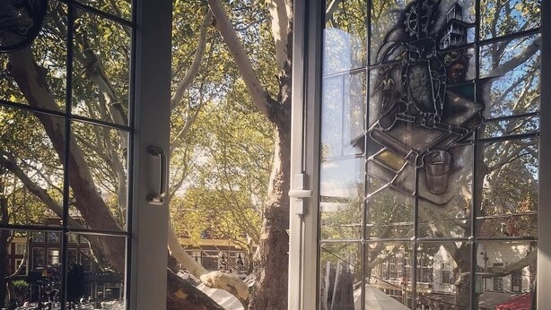 A view through large windows of an outdoor seating area with tables and chairs, surrounded by trees. - Hotel de Koophandel (Delft)