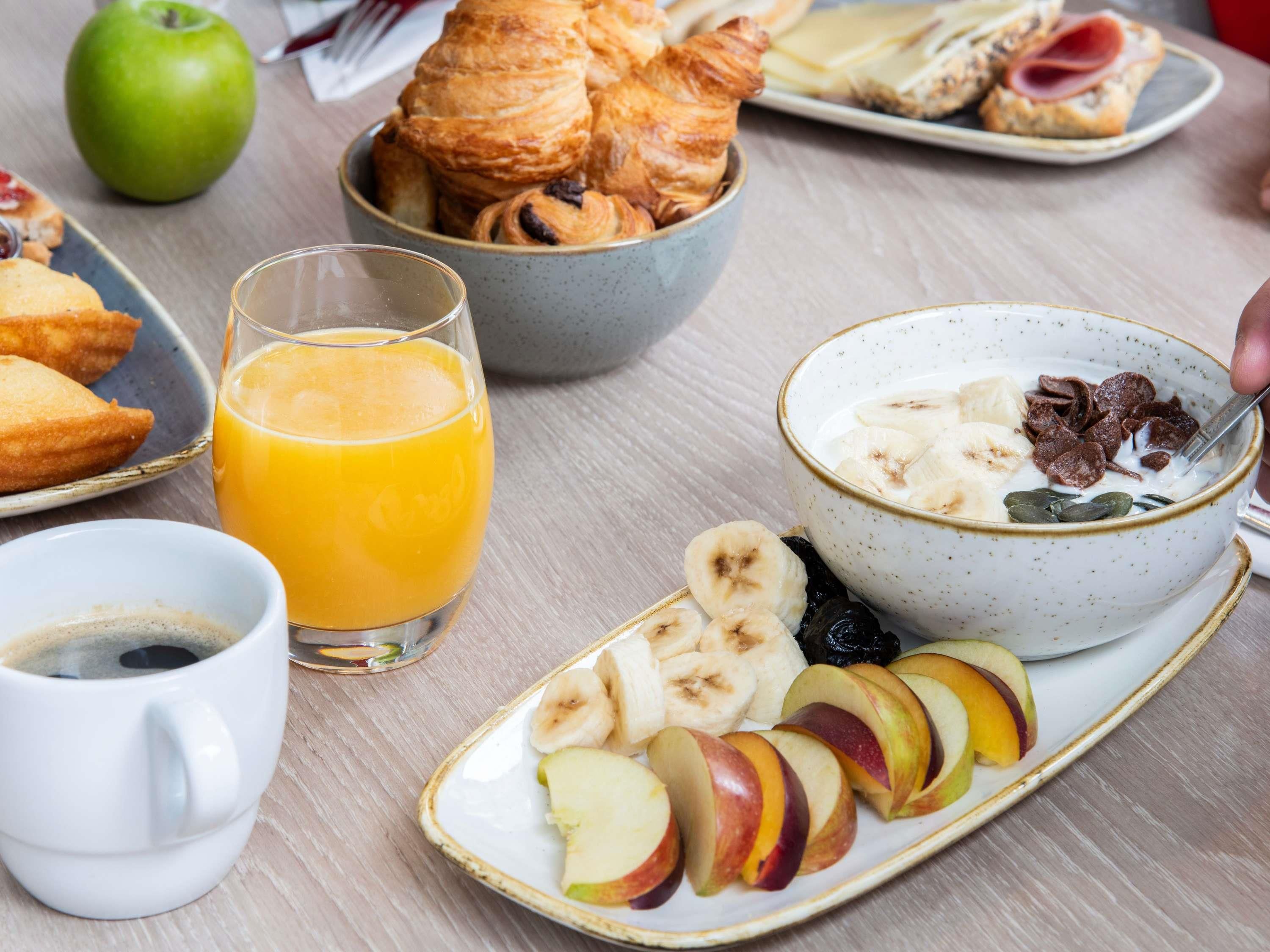 A breakfast setting with croissants, a bowl of cereal with fruit, a glass of orange juice, and a plate of sliced apples.