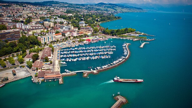 Vista aérea de una ciudad costera con un puerto deportivo, edificios y un mar azul claro.