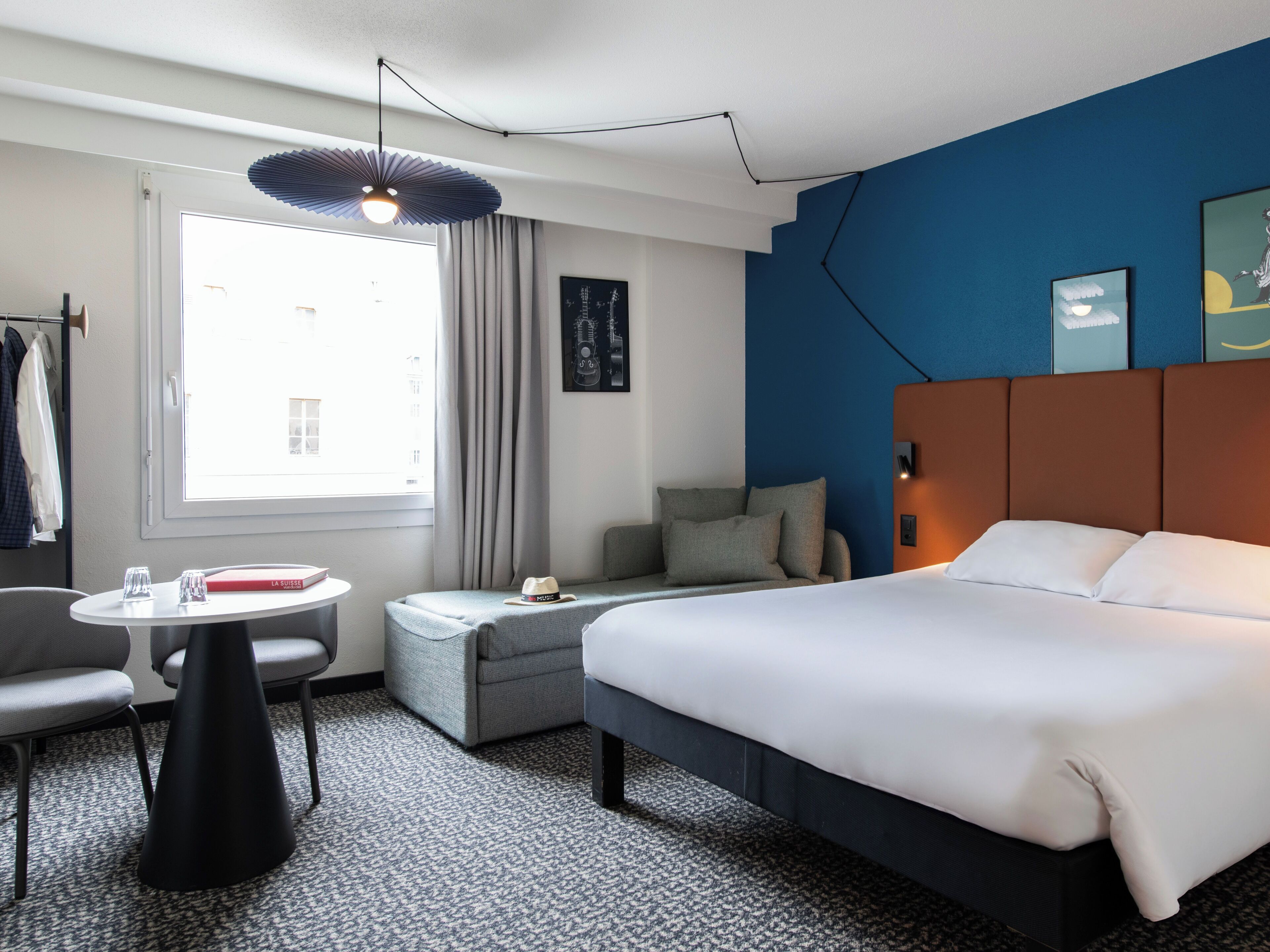 Hotellihuoneessa on sänky, sohva, pieni pöytä ja tuoli. Siellä on myös ikkuna, jossa on verhot, seinäkello ja taulu.