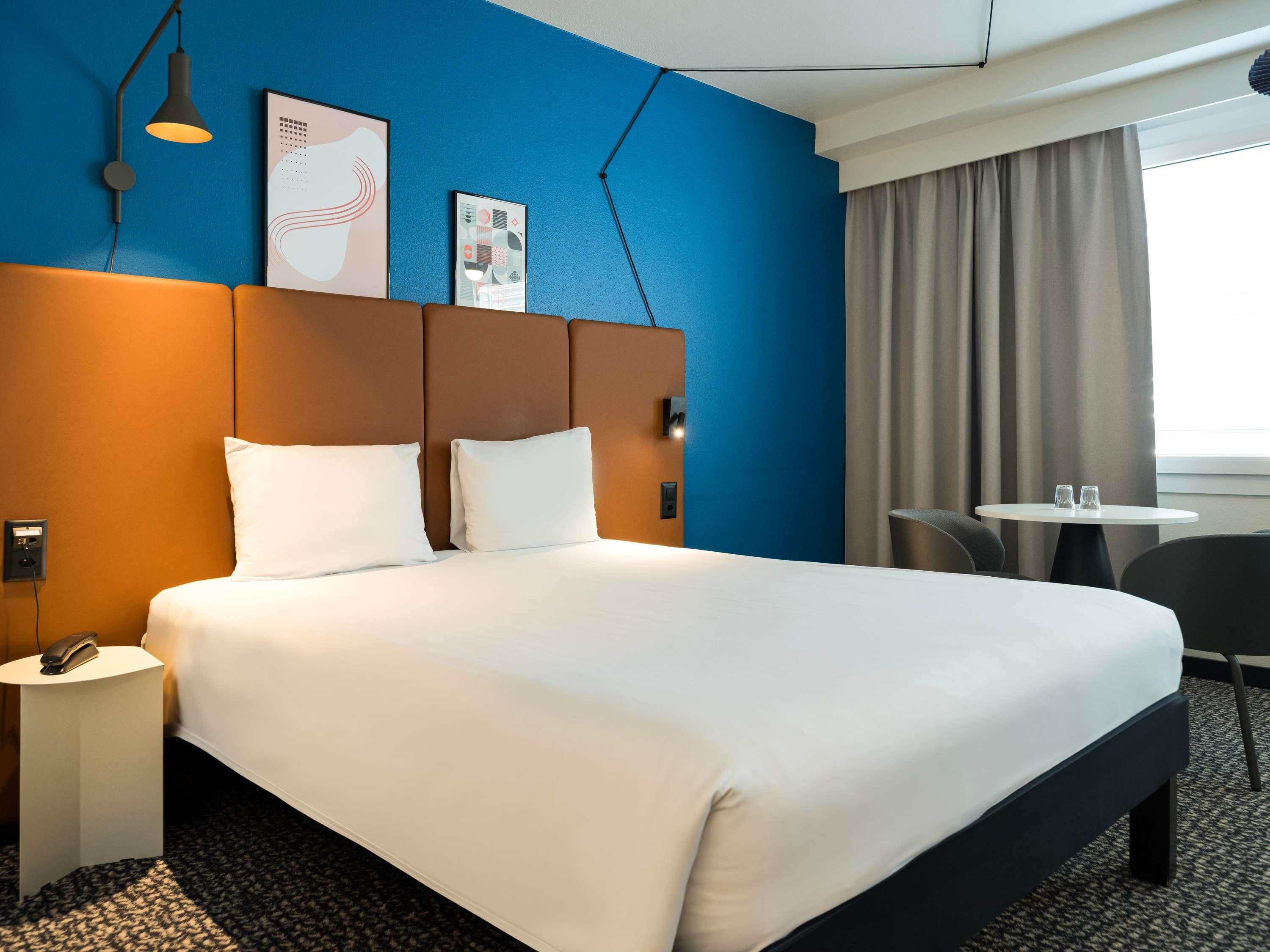 A hotel room with a large bed, a bedside table with a phone, a small table with two glasses, and a chair.