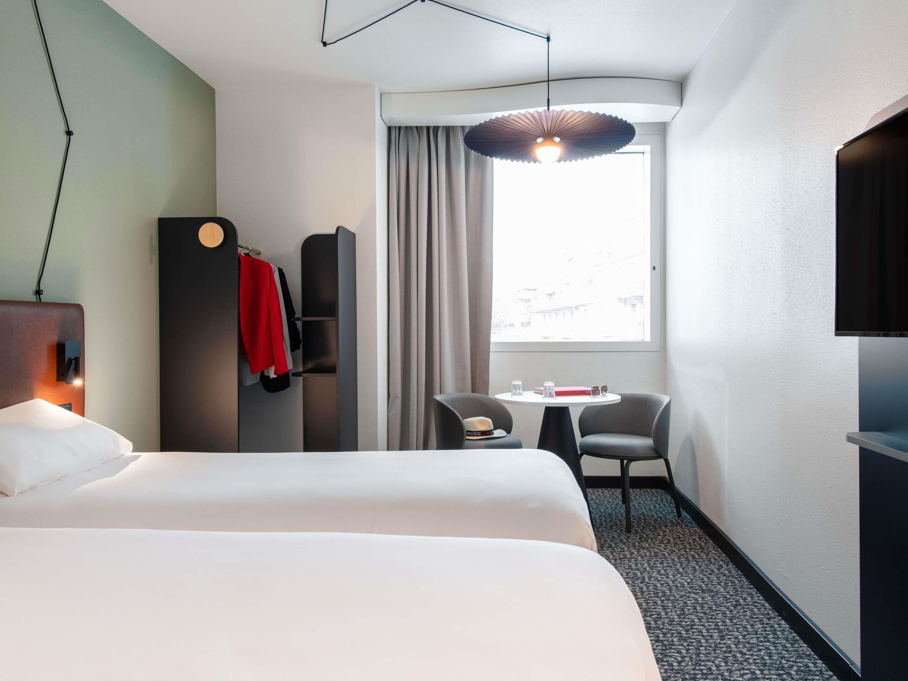 A hotel room with a bed, a desk with two chairs, a TV, and a window with curtains.