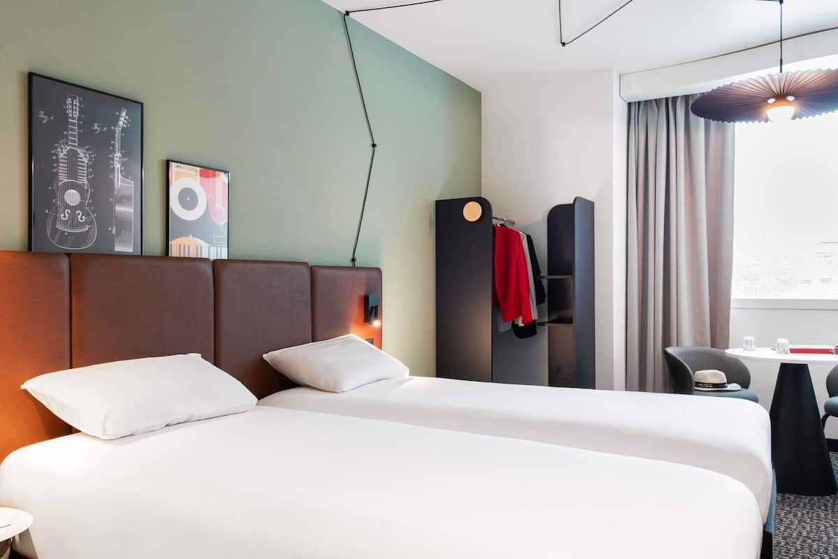 Een hotelkamer met een groot bed, een bureau en een stoel. Twee ingelijste foto’s hangen aan de muur, er is een klok en een raam met gordijnen.