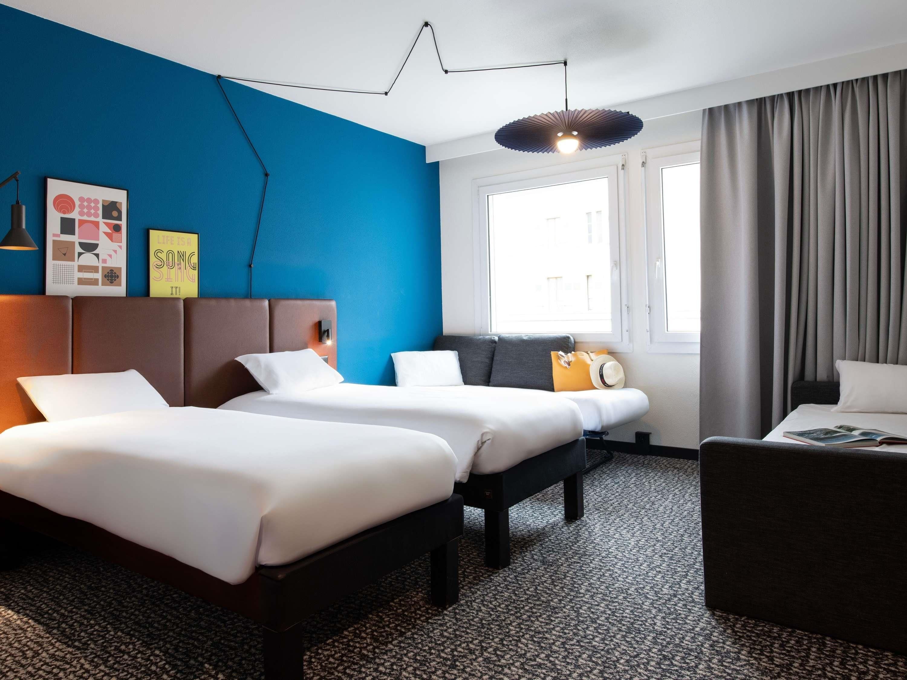 A hotel room with two beds, a sofa, a desk, and a large window with curtains.