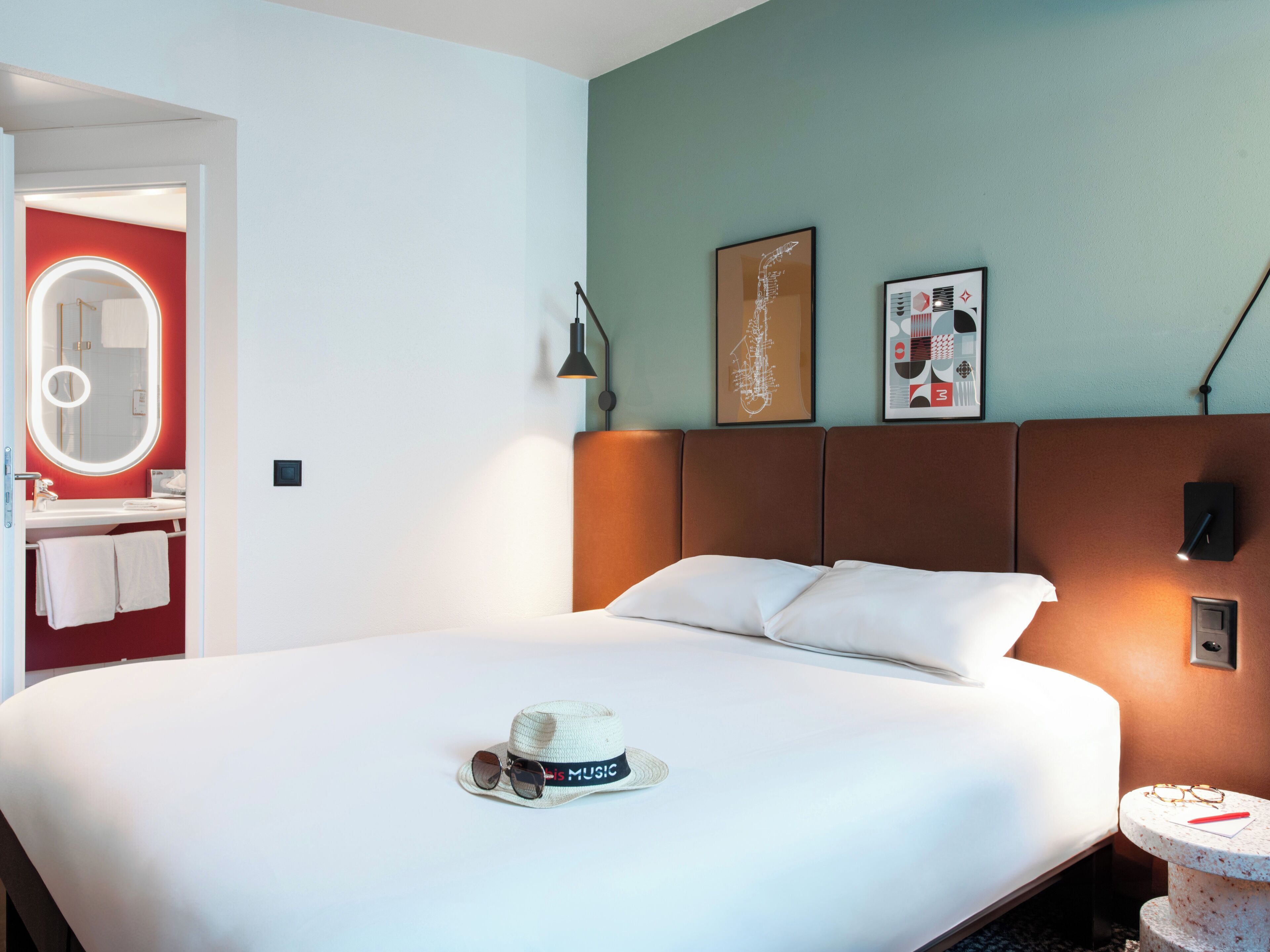 Una habitación de hotel con una cama, un espejo redondo con luz, una mesita de noche con lámpara y una obra de arte en la pared.