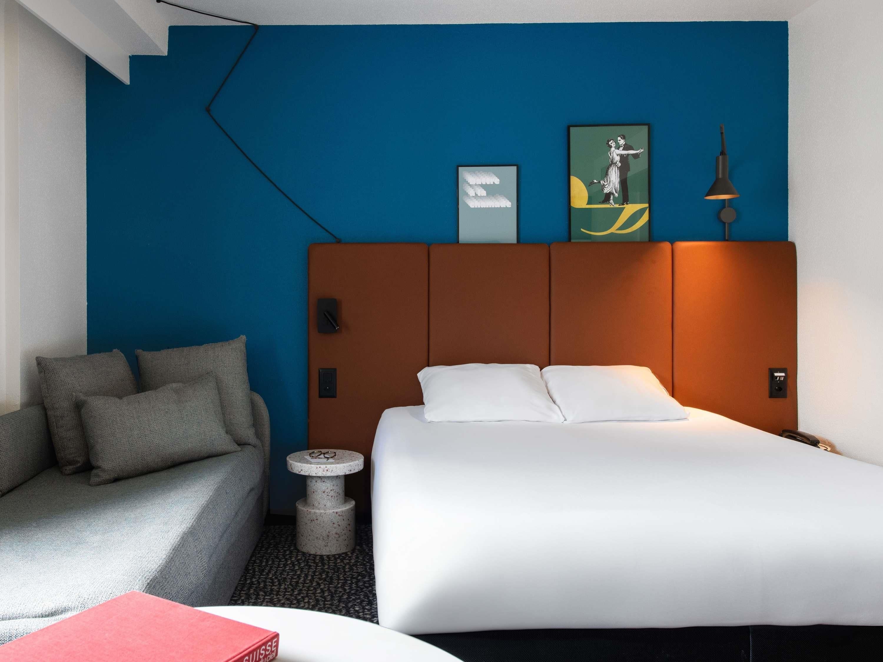 A hotel room with a bed, a sofa, a small round table, and a wall with artwork.