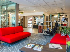 A modern hotel lobby with a red sofa, a black chair, and a coffee table with magazines. There are bookshelves with books and decorative items.