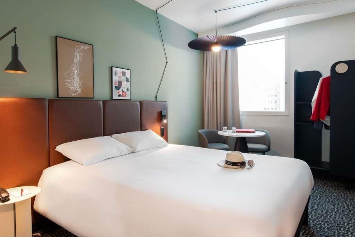 A hotel room with a large bed, a desk with a chair, a round table, a window with curtains, and a coat rack.