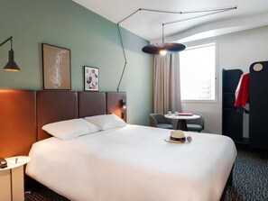 A hotel room with a large bed, a desk with a chair, a round table, a window with curtains, and a coat rack. - ibis Lausanne Centre (Lausanne)