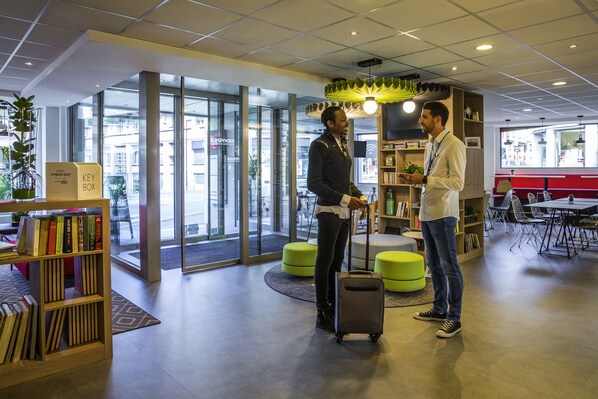 Een moderne hotellobby met een glazen ingang, een houten boekenkast en een zithoek met groene poefs.