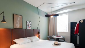 Habitación de hotel con una cama grande, mesita de noche, lámpara, obra de arte, ventana con cortinas y perchero.