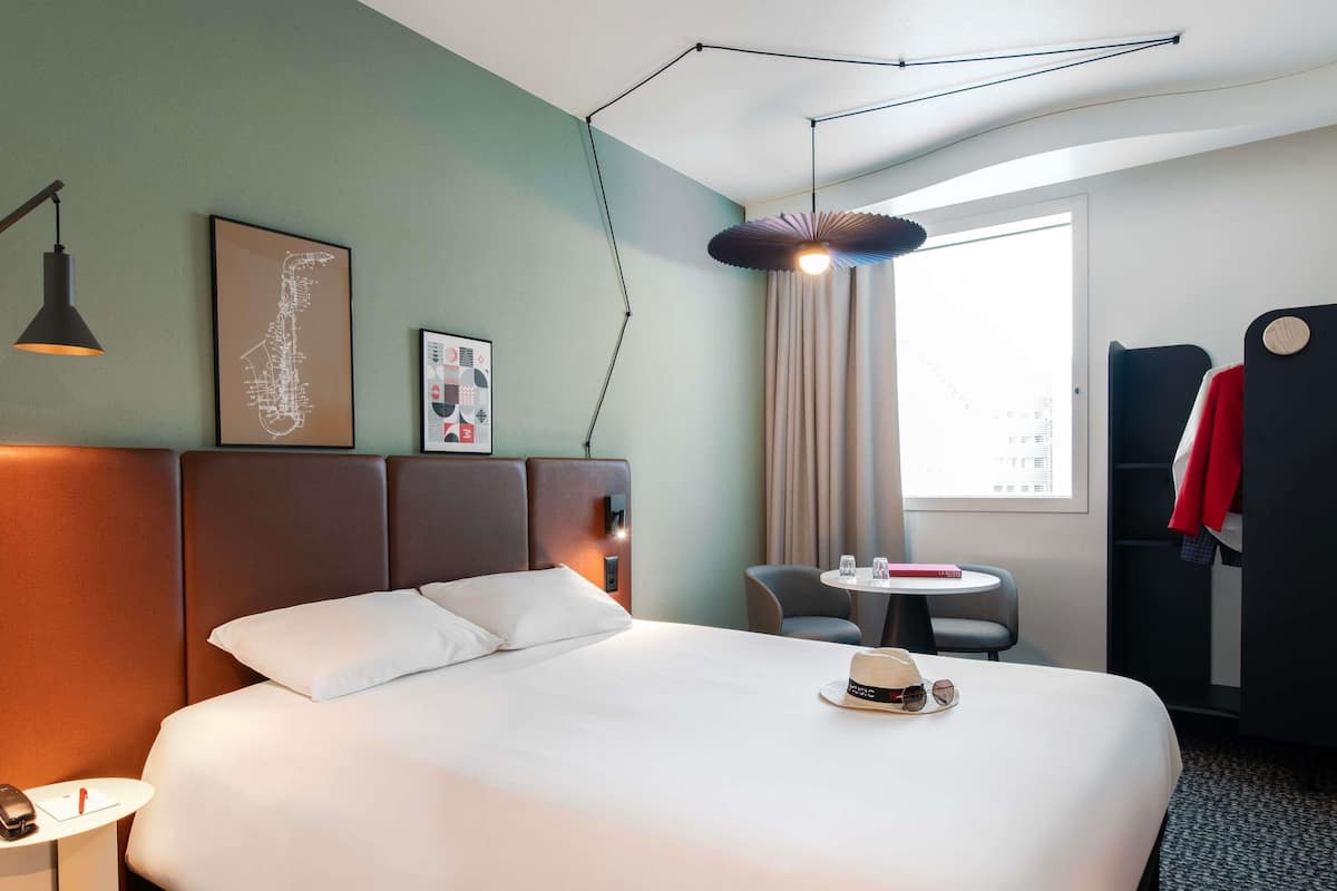 Een hotelkamer met een groot bed, nachtkastje, lamp, kunstwerk, raam met gordijnen en een kapstok.