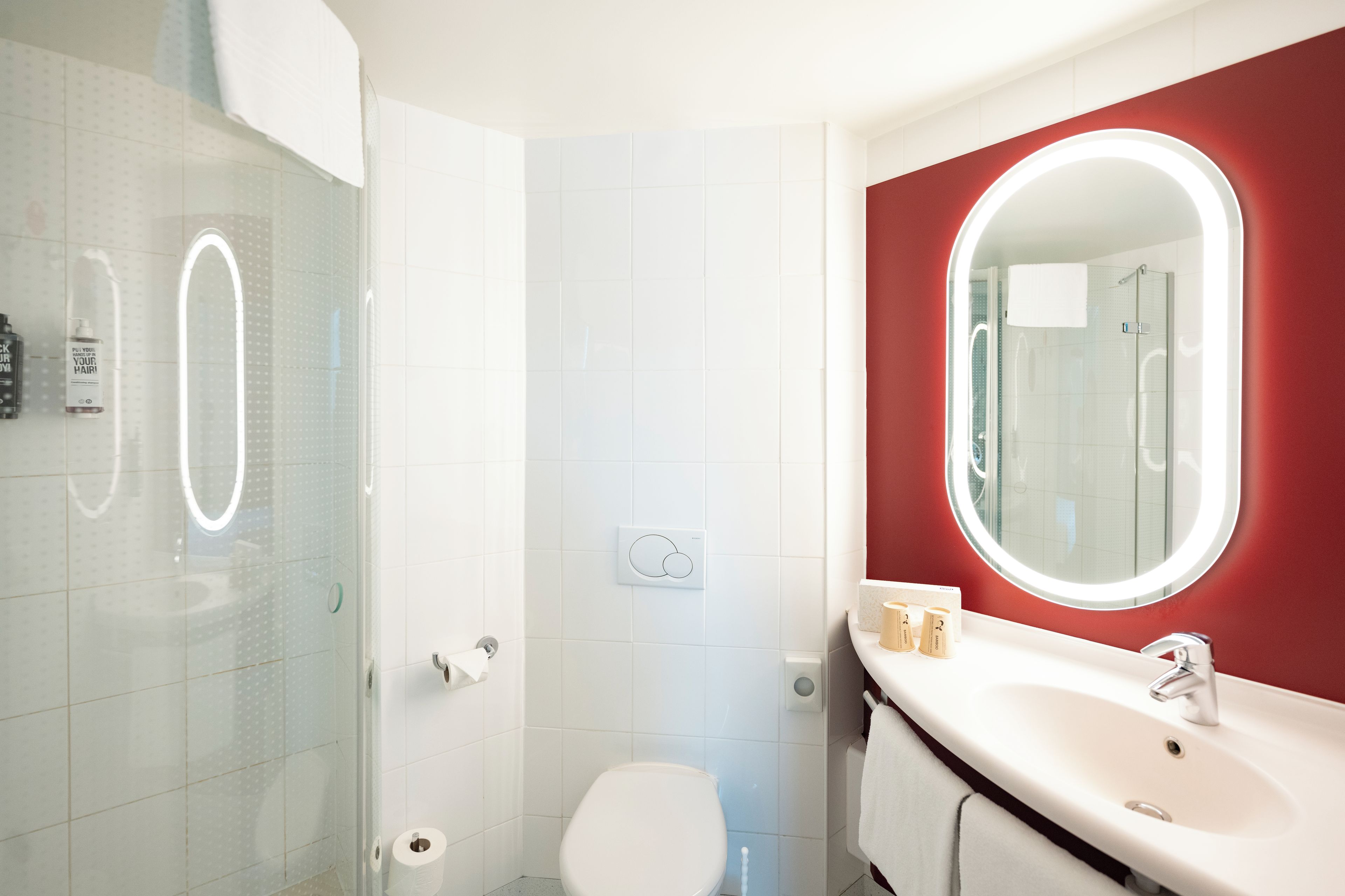 A bathroom with a glass shower enclosure, a white toilet, a vanity with a mirror, and a red accent wall.