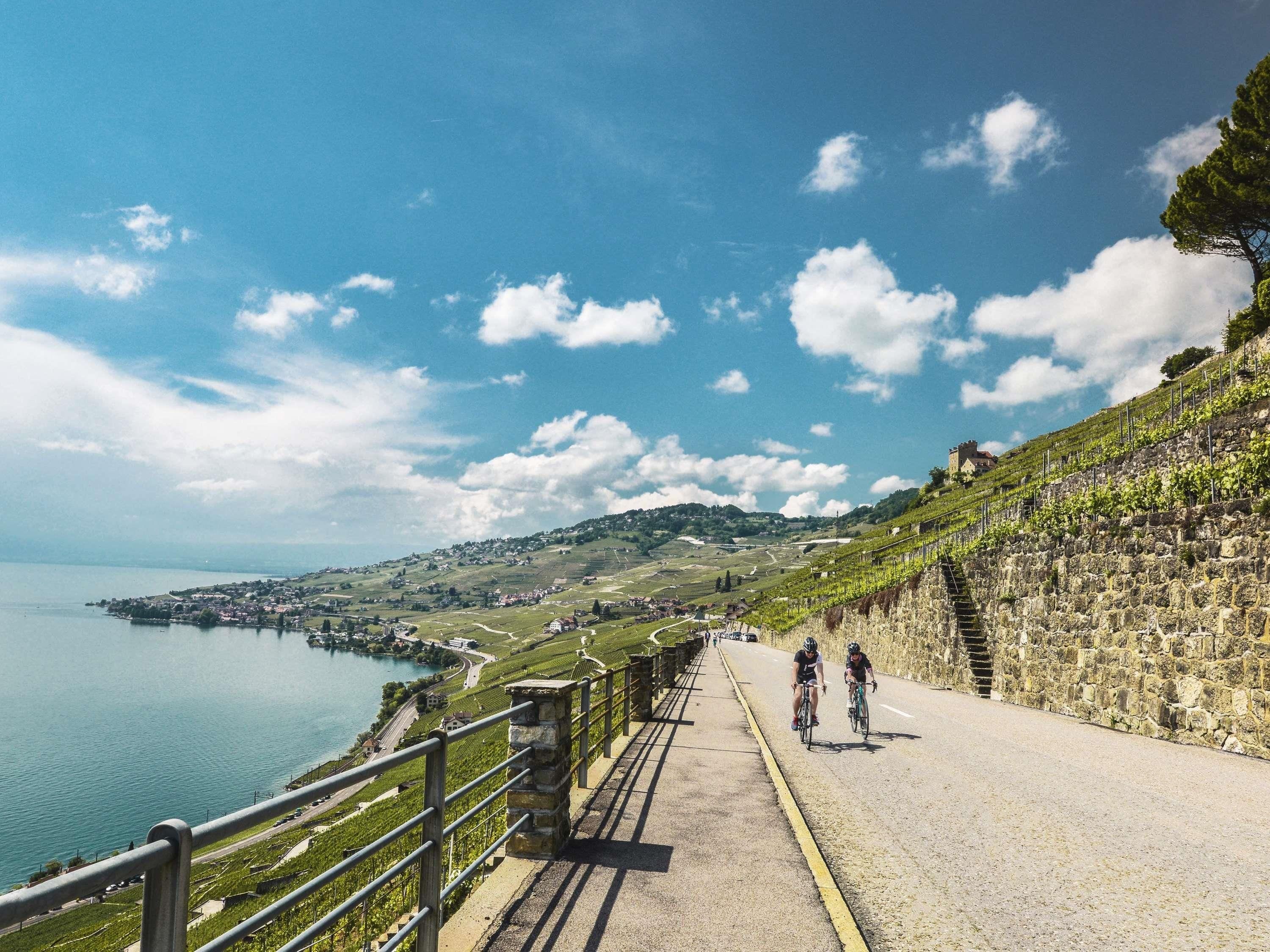 A paved pathway alongside a body of water with a stone wall on one side and a grassy hillside on the other.