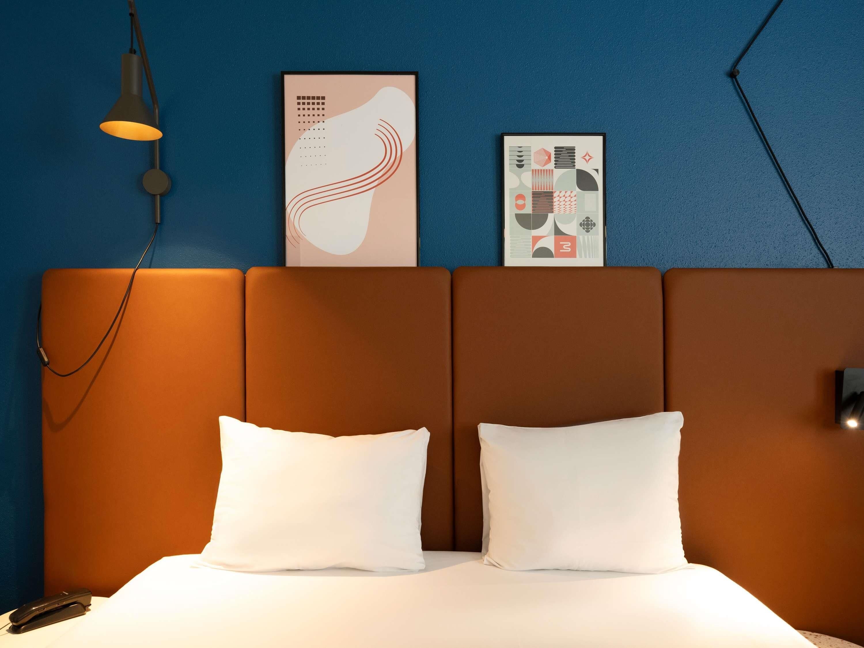 Habitación de hotel con cama, dos almohadas, una mesita de noche, lámpara y dos obras de arte en la pared.