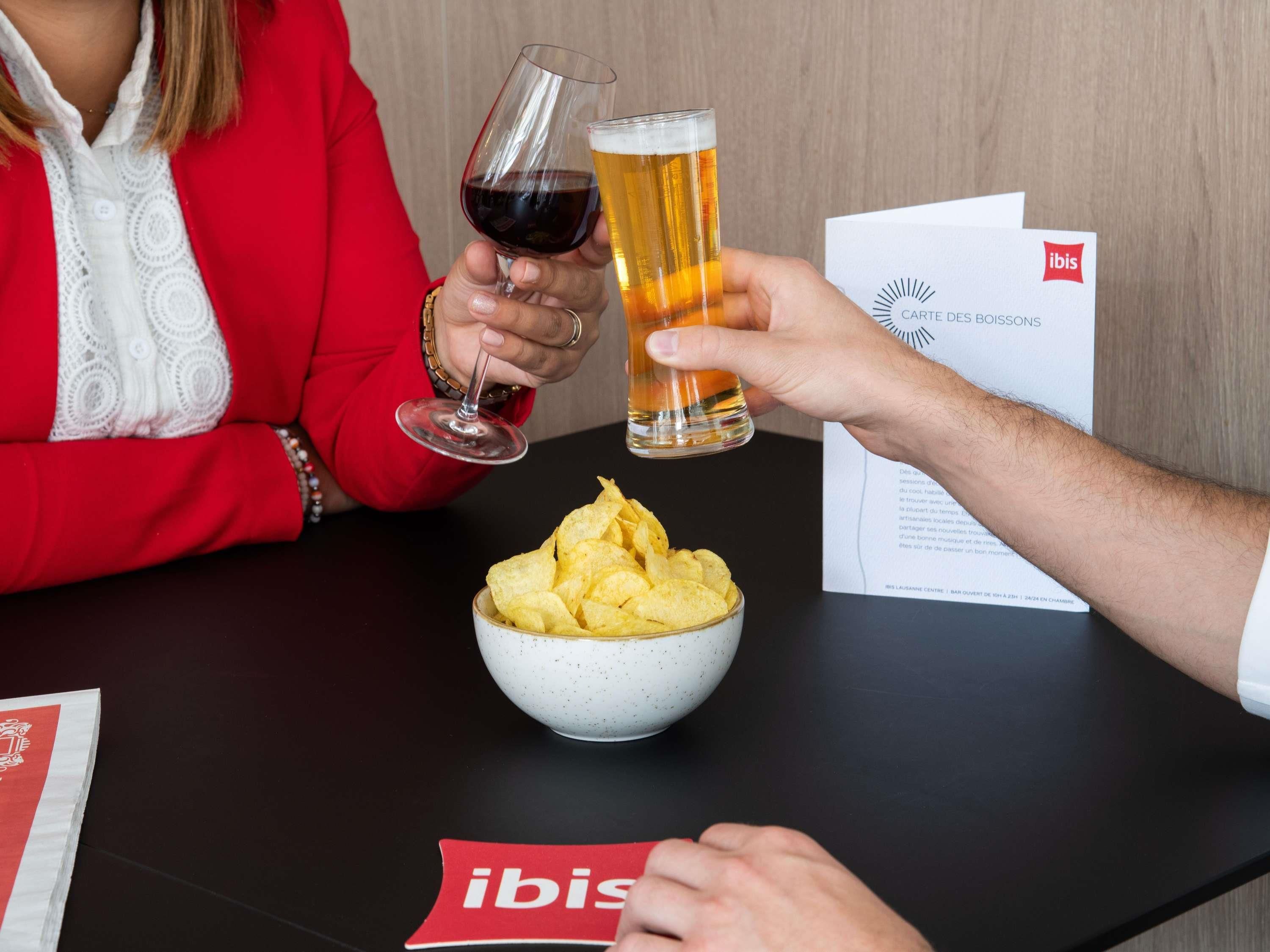 A person in a red jacket holding a wine glass and a beer glass, with a bowl of chips on the table.