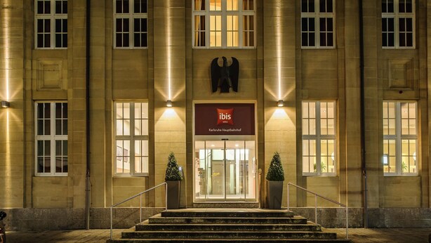 A hotel entrance with a prominent 'ibis HOTEL' sign, a double door entrance, and two potted plants on the steps. - ibis Karlsruhe Hauptbahnhof (Karlsruhe)