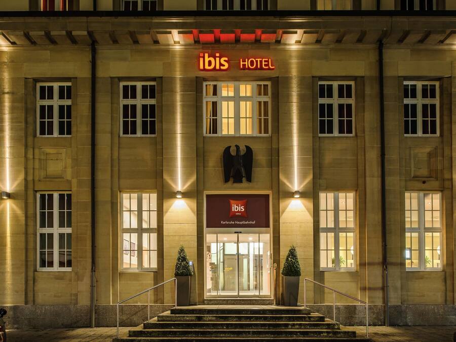 A hotel entrance with a prominent 'ibis HOTEL' sign, a double door entrance, and two potted plants on the steps.