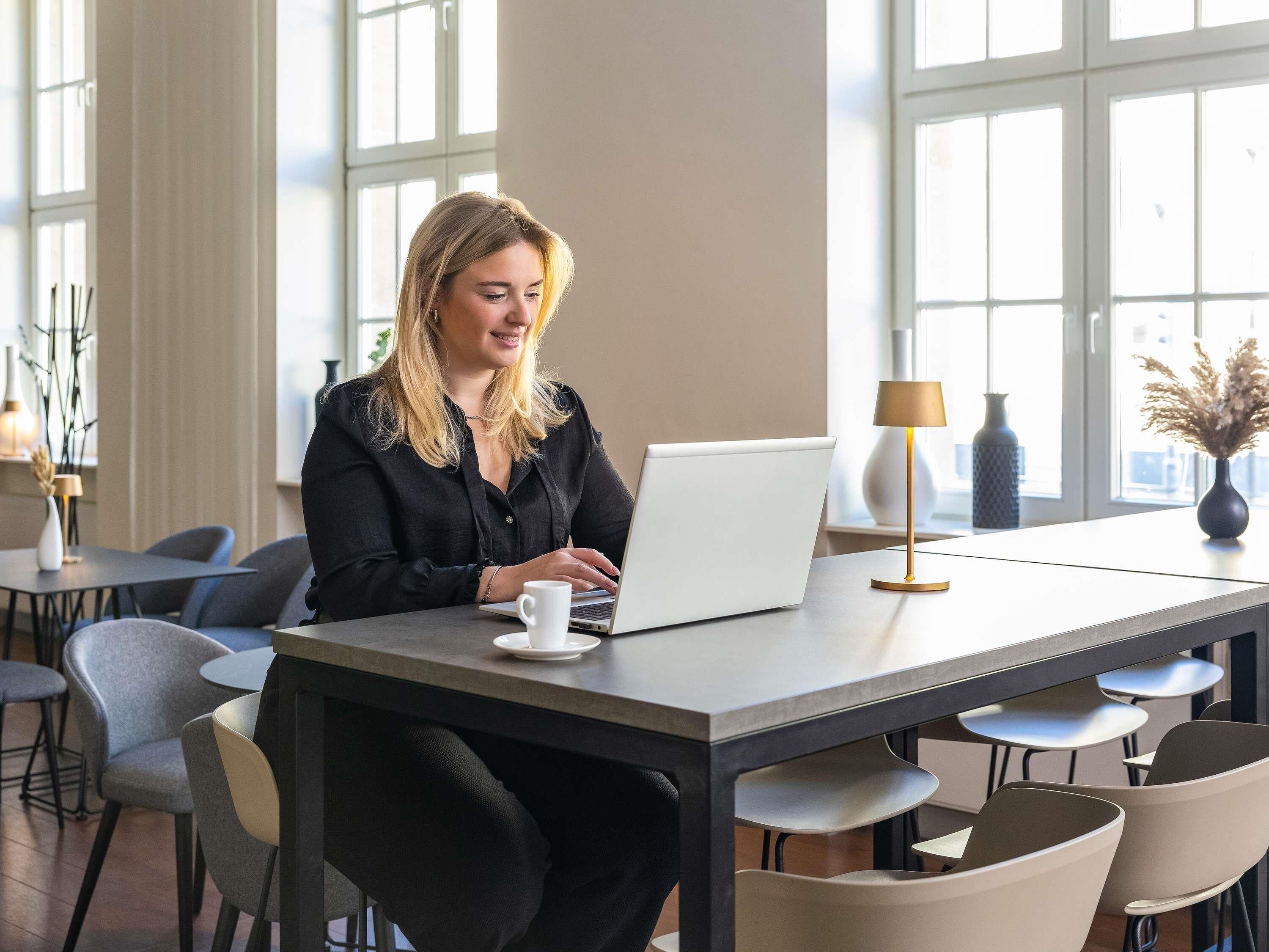 Iemand werkt op een laptop in een moderne kantooromgeving, met meerdere stoelen, een tafel en een lamp.