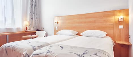 Habitación de hotel con dos camas, cabecero de madera, escritorio con lámpara y una mesita.