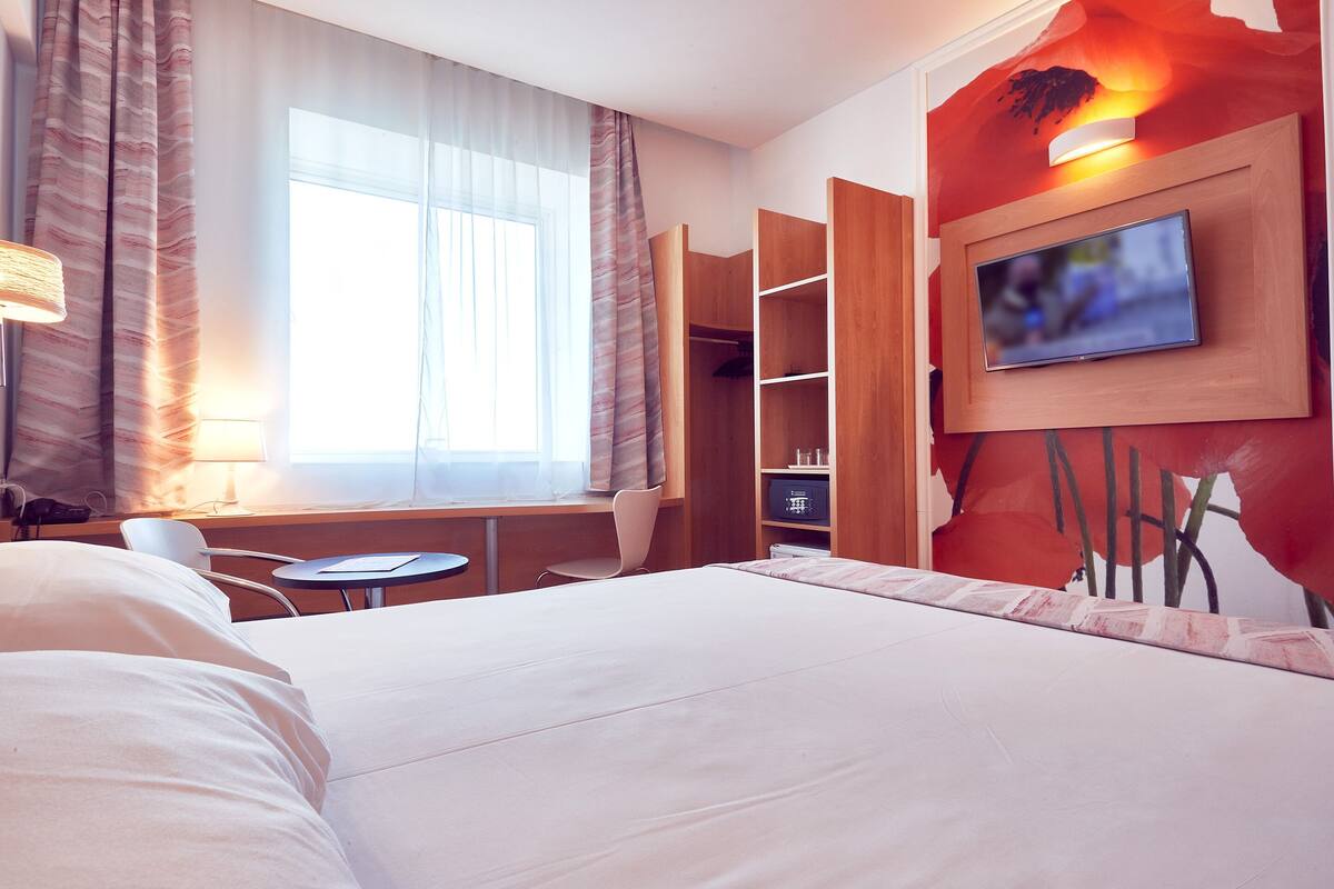 A hotel room with a large bed, a desk with a chair, a TV mounted on the wall, and a wardrobe.