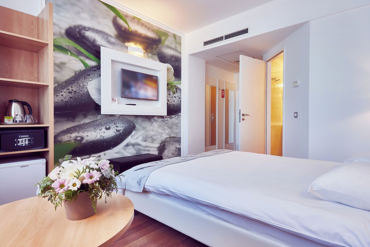 Habitación de hotel con cama, televisión montada en la pared, una mesita con un arreglo floral y una zona de kitchenette.