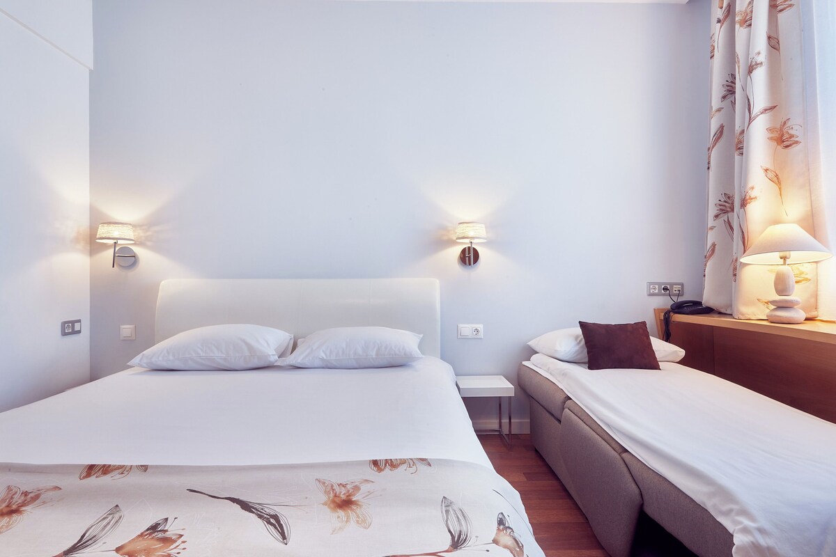 Una habitación de hotel con cama, mesita de noche, lámpara y una pequeña mesita auxiliar.
