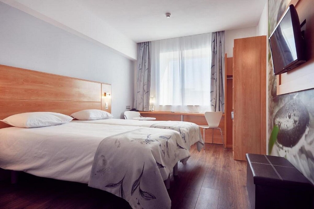 Habitación de hotel con una cama grande, un escritorio, una silla, un televisor y una ventana con cortinas.