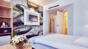 Habitación de hotel con cama, televisión montada en la pared, una mesita con un arreglo floral y una zona de kitchenette.