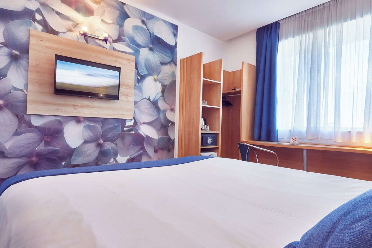 Habitación de hotel con cama, televisión montada en un panel de madera, armario y escritorio con silla.