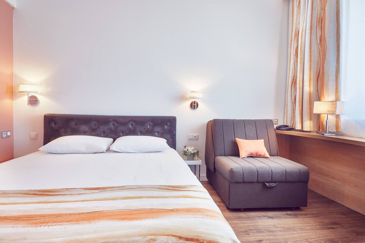 A hotel room with a large bed, a grey armchair, and a small table with a vase of flowers.