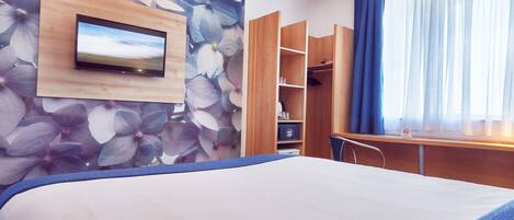 A hotel room with a bed, a TV mounted on a wooden panel, a wardrobe, and a desk with a chair.