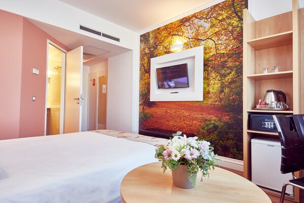 A hotel room with a bed, a TV mounted on a wall with a nature mural, a small desk with a kettle, and a chair. - MyContinental Sibiu (Sibiu)