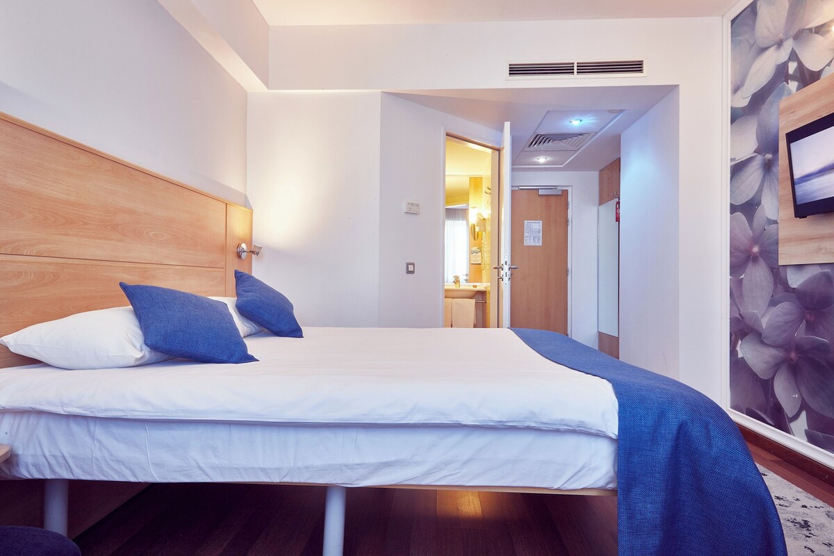 Una habitación de hotel con una cama, cabecera de madera, manta azul y televisión instalada en la pared.