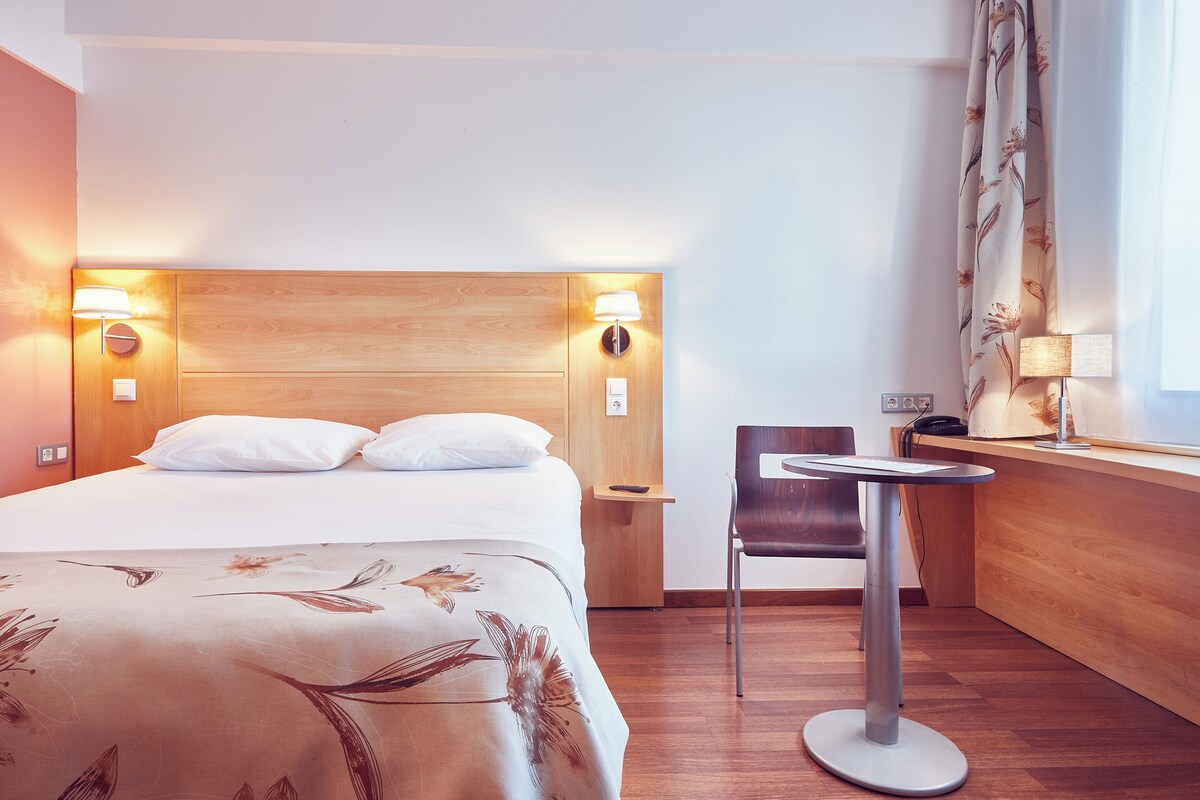 Habitación de hotel con cabecera de madera, cama con ropa blanca, una mesita con una silla y una ventana con cortinas.