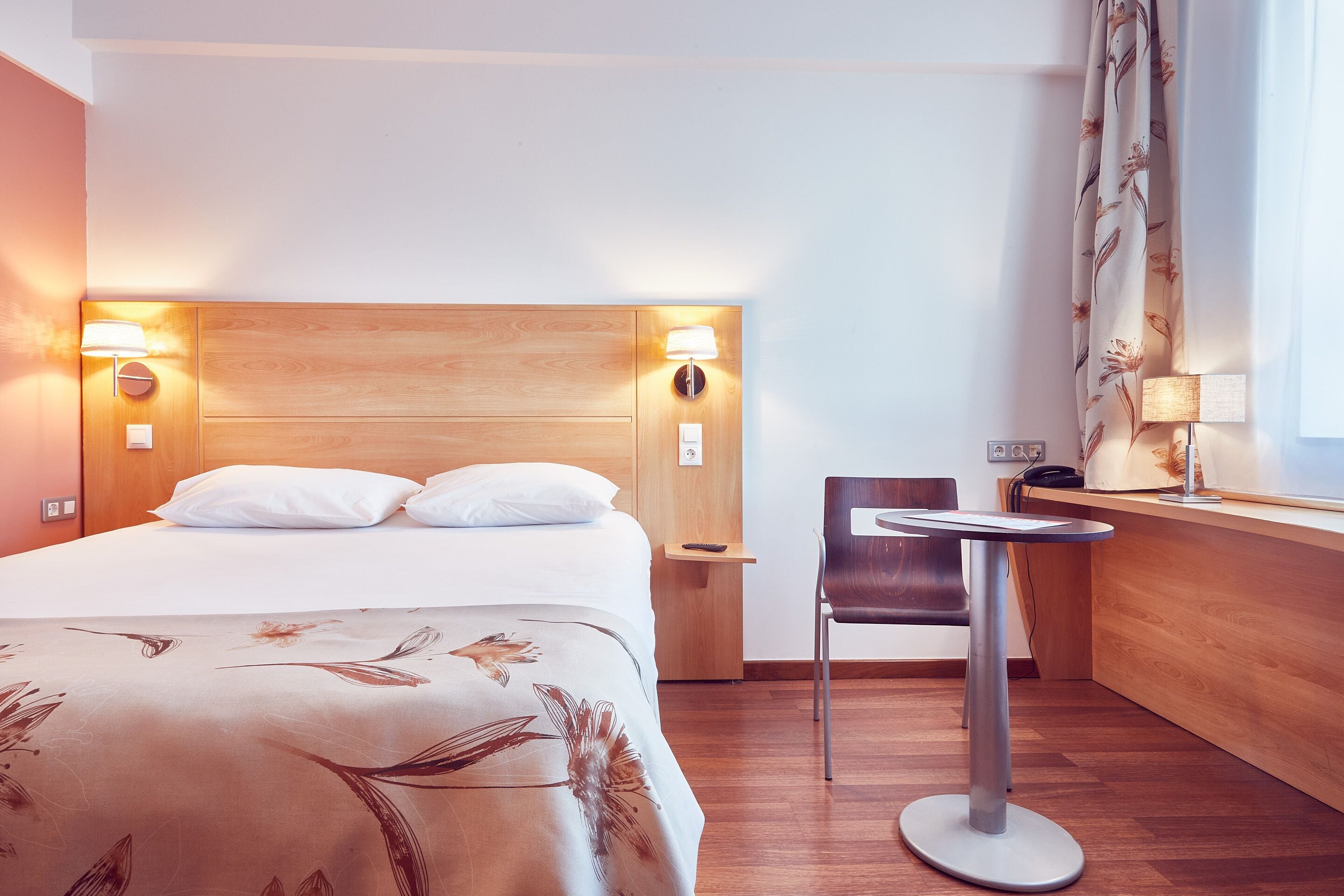 A hotel room with a wooden headboard, a bed with white linens, a small table with a chair, and a window with curtains.