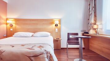 Ein Hotelzimmer mit einem hölzernen Kopfteil, einem Bett mit weißer Bettwäsche, einem kleinen Tisch mit einem Stuhl und einem Fenster mit Vorhängen.