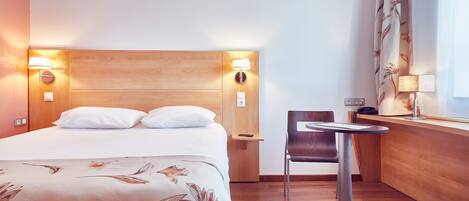 A hotel room with a wooden headboard, a bed with white linens, a small table with a chair, and a window with curtains.