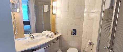 A compact bathroom with a white sink, mirror, and toilet. There is a towel rack with towels, a trash bin, and a shower area with a glass door.