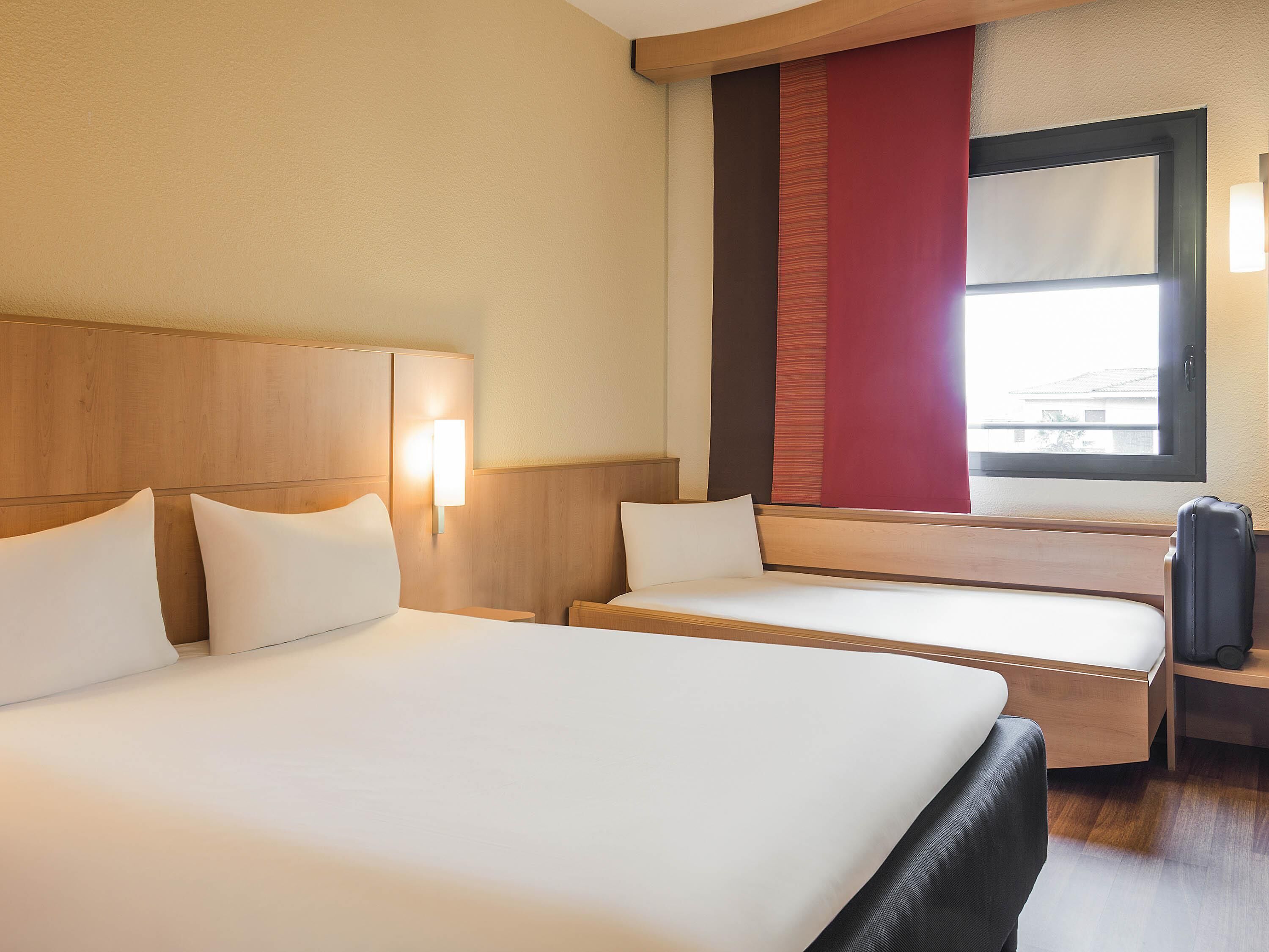 Una camera d'albergo con due letti, una testiera in legno, una lampada da comodino, una finestra con vista e una valigia su un comodino.