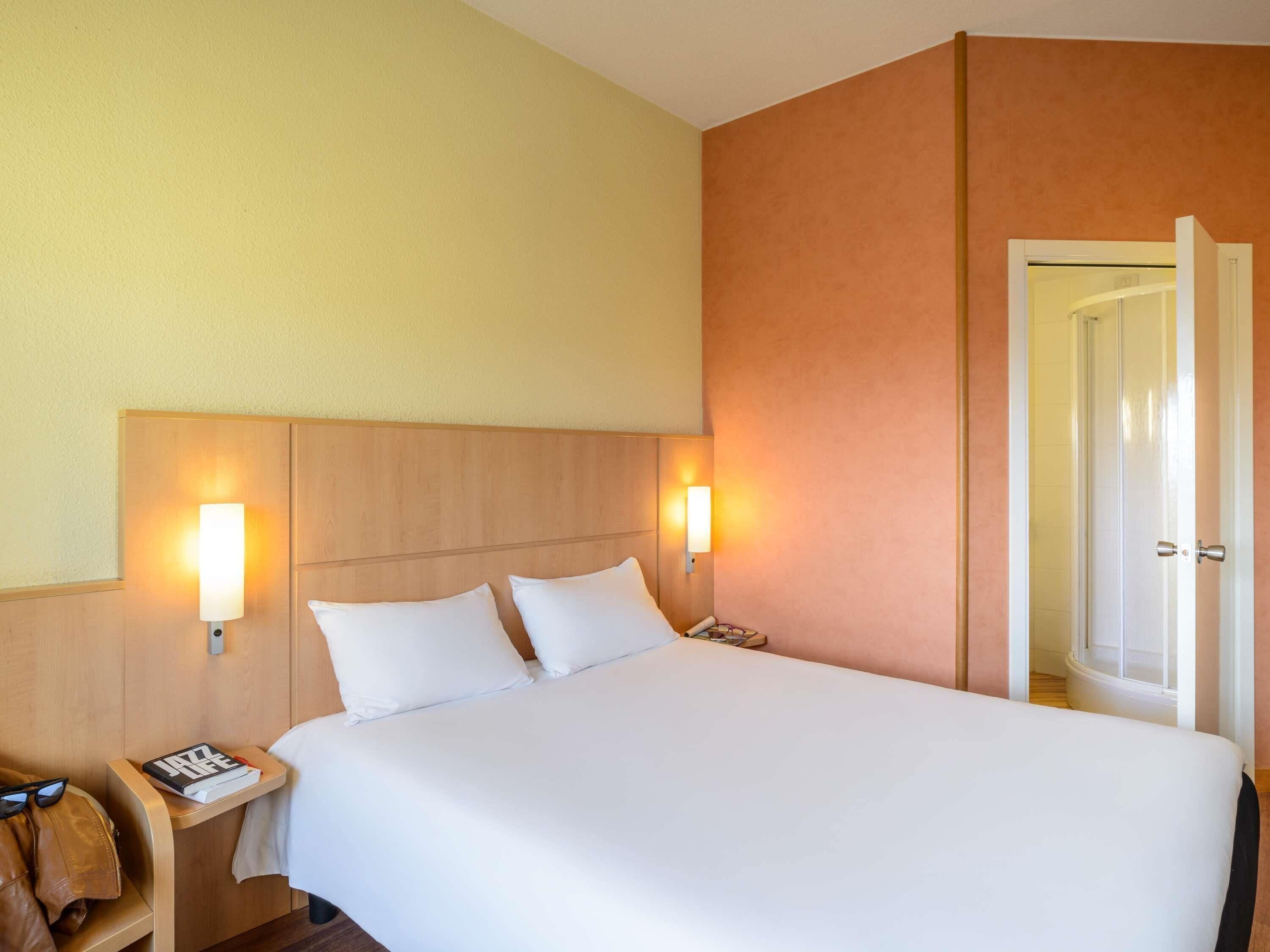 Une chambre d’hôtel avec un lit, deux lampes de chevet, une table de chevet avec un livre et des lunettes, et une porte donnant accès à la salle de bain.
