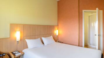 Een hotelkamer met een bed, twee bedlampen, een nachtkastje met een boek en brillen, en een deur naar de badkamer.