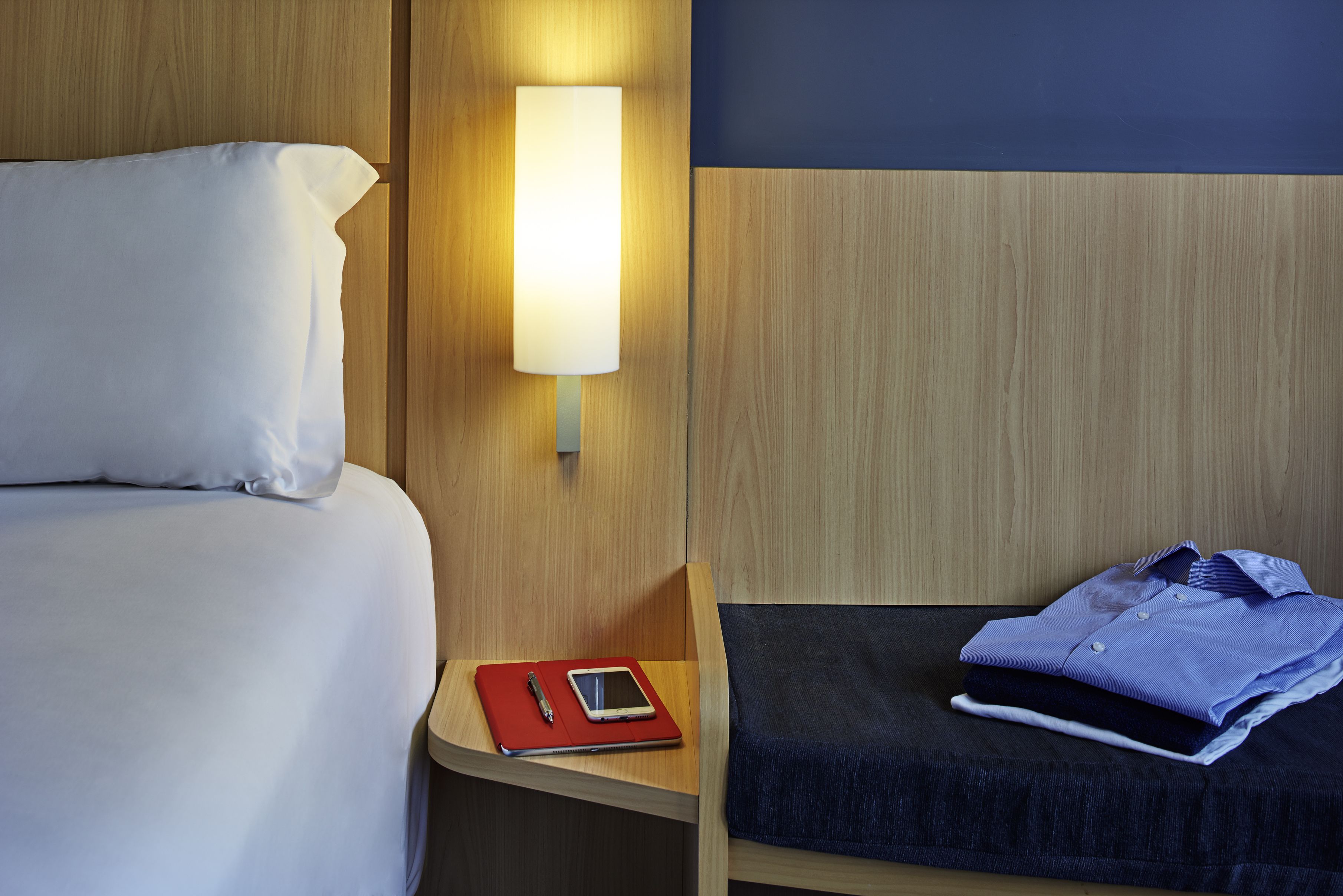 Una cama bien hecha con sábanas blancas, una mesita de noche con una carpeta roja y una tableta, y una camisa azul doblada cuidadosamente sobre un almohadón azul oscuro.