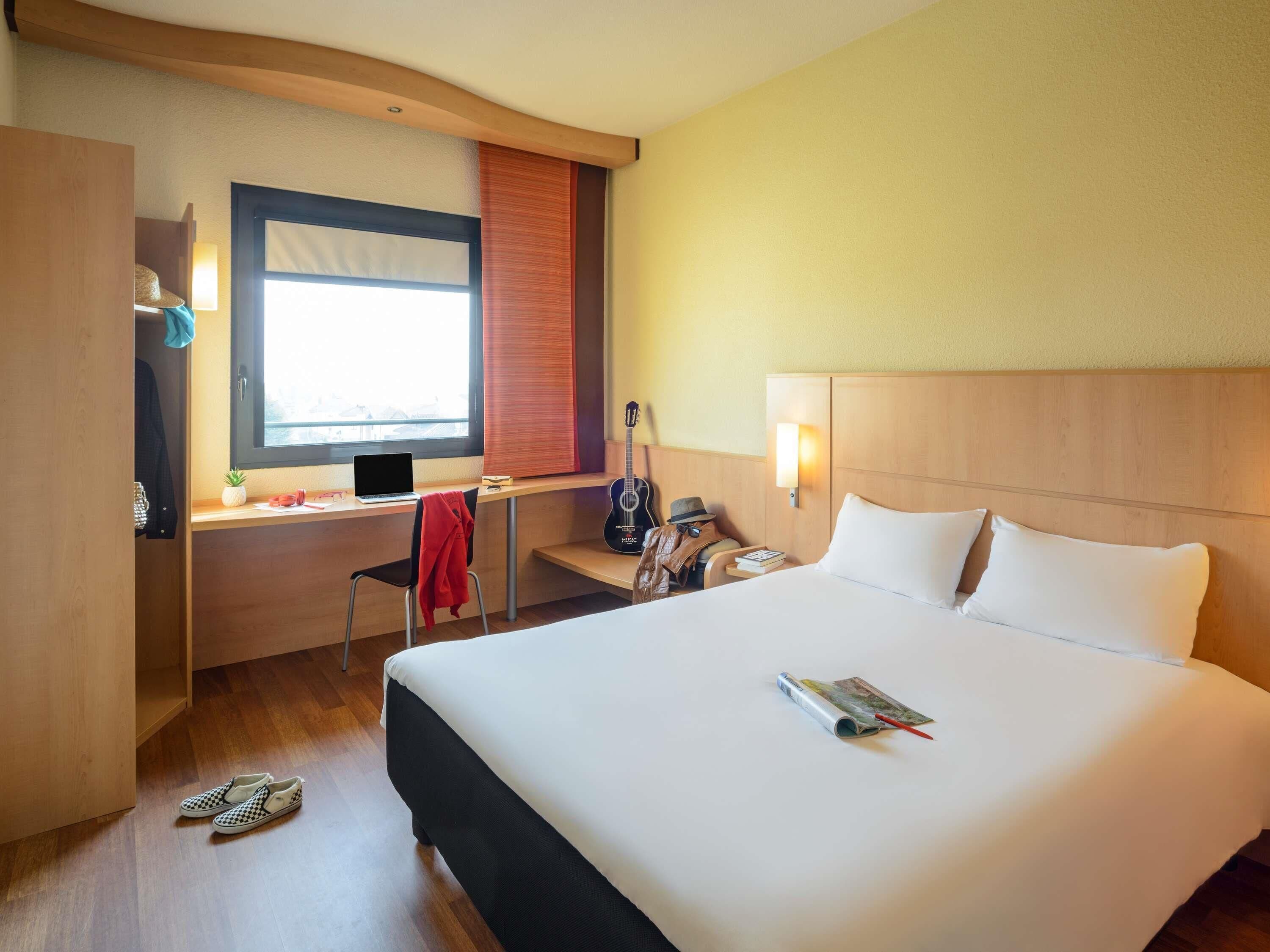 Quarto de hotel com uma cama, uma mesa com um laptop, um violão e uma cadeira.