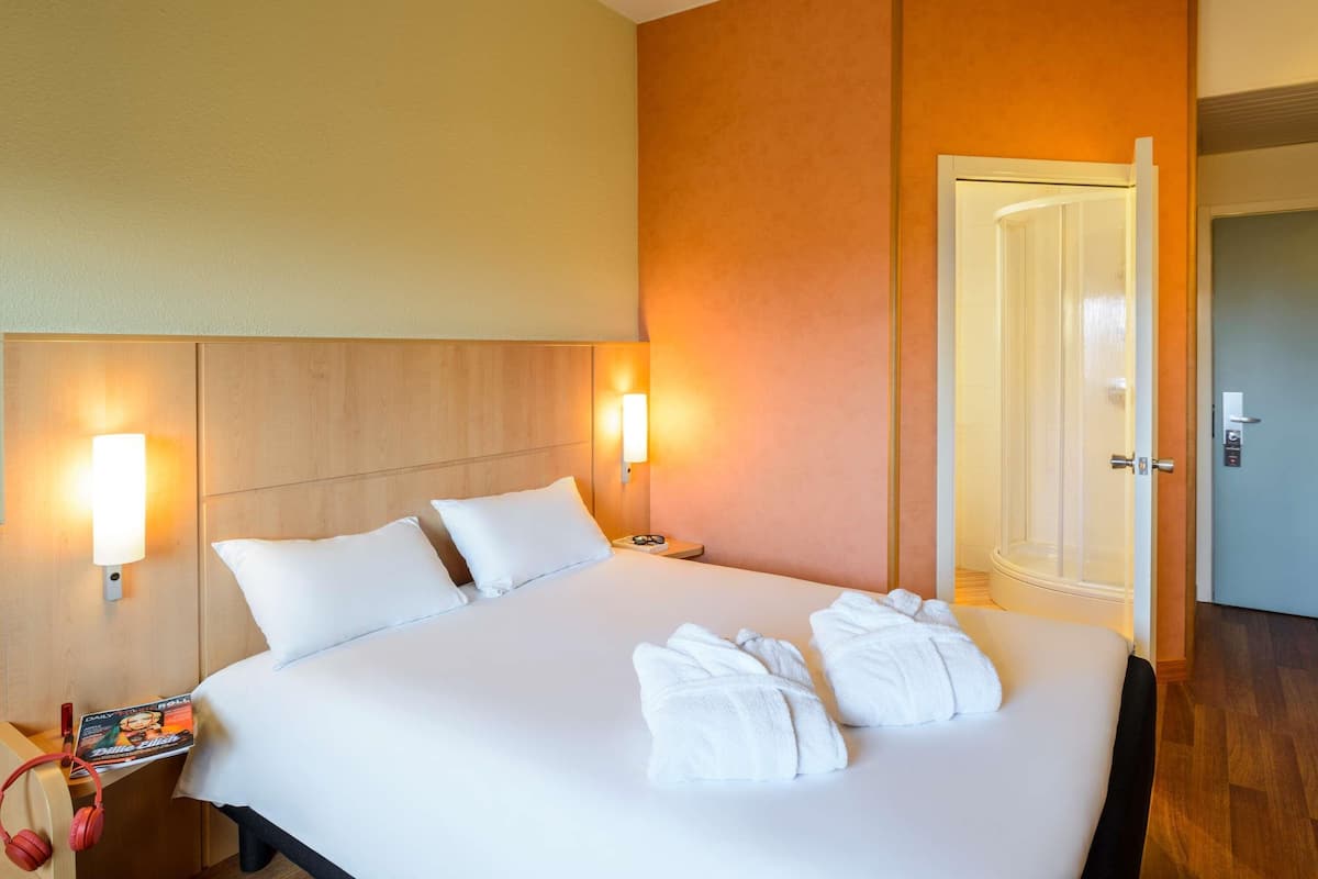 Habitación de hotel con una cama grande, dos lámparas de noche, una mesita de noche con una revista y auriculares, y una puerta a la derecha.