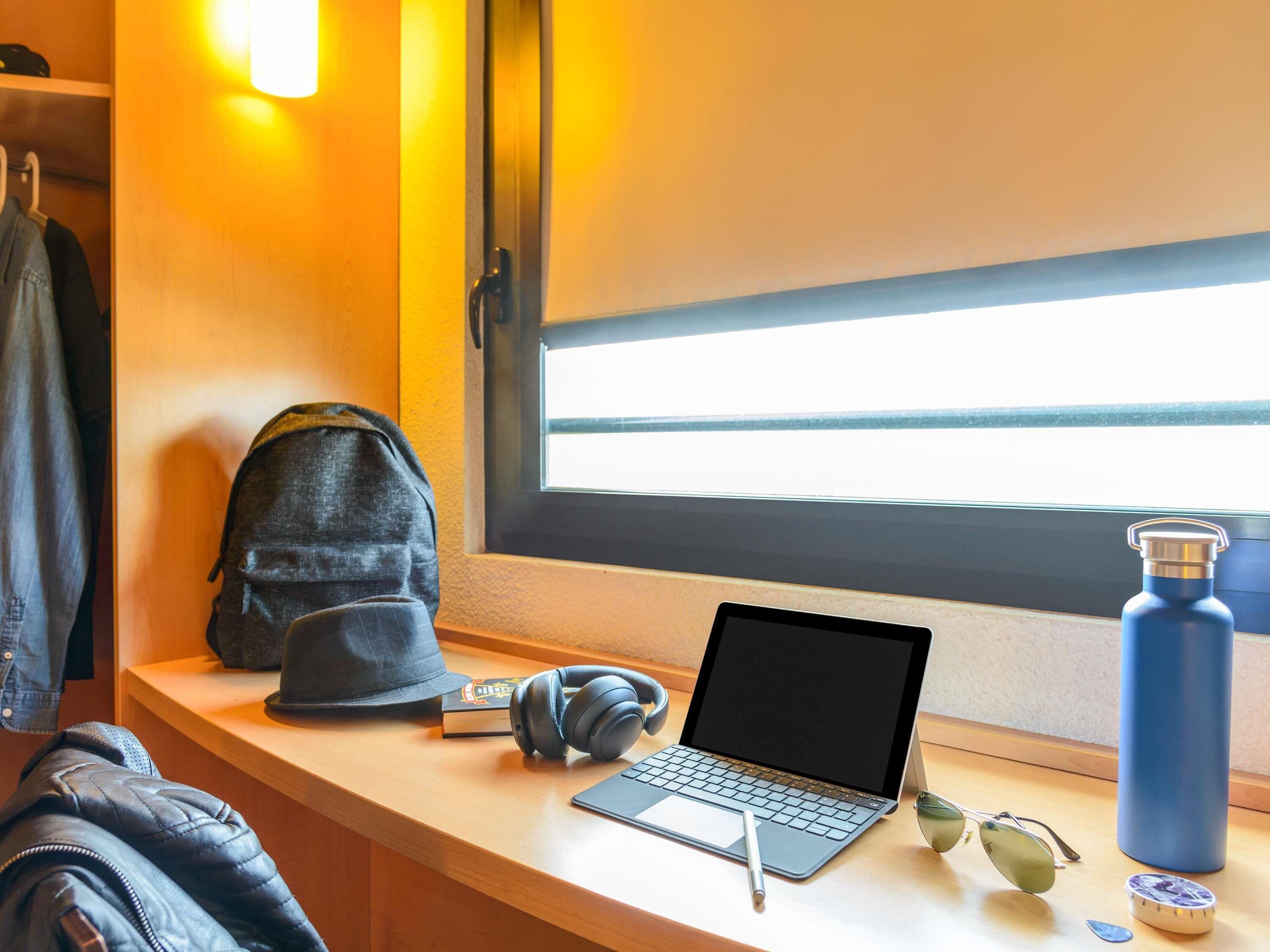 A laptop on a desk with a window, a backpack, headphones, a pen, a pair of sunglasses, and a water bottle.