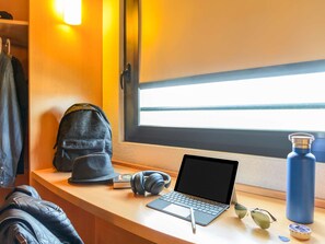 Una computadora portátil sobre un escritorio con una ventana, una mochila, auriculares, un bolígrafo, unas gafas de sol y una botella de agua.
