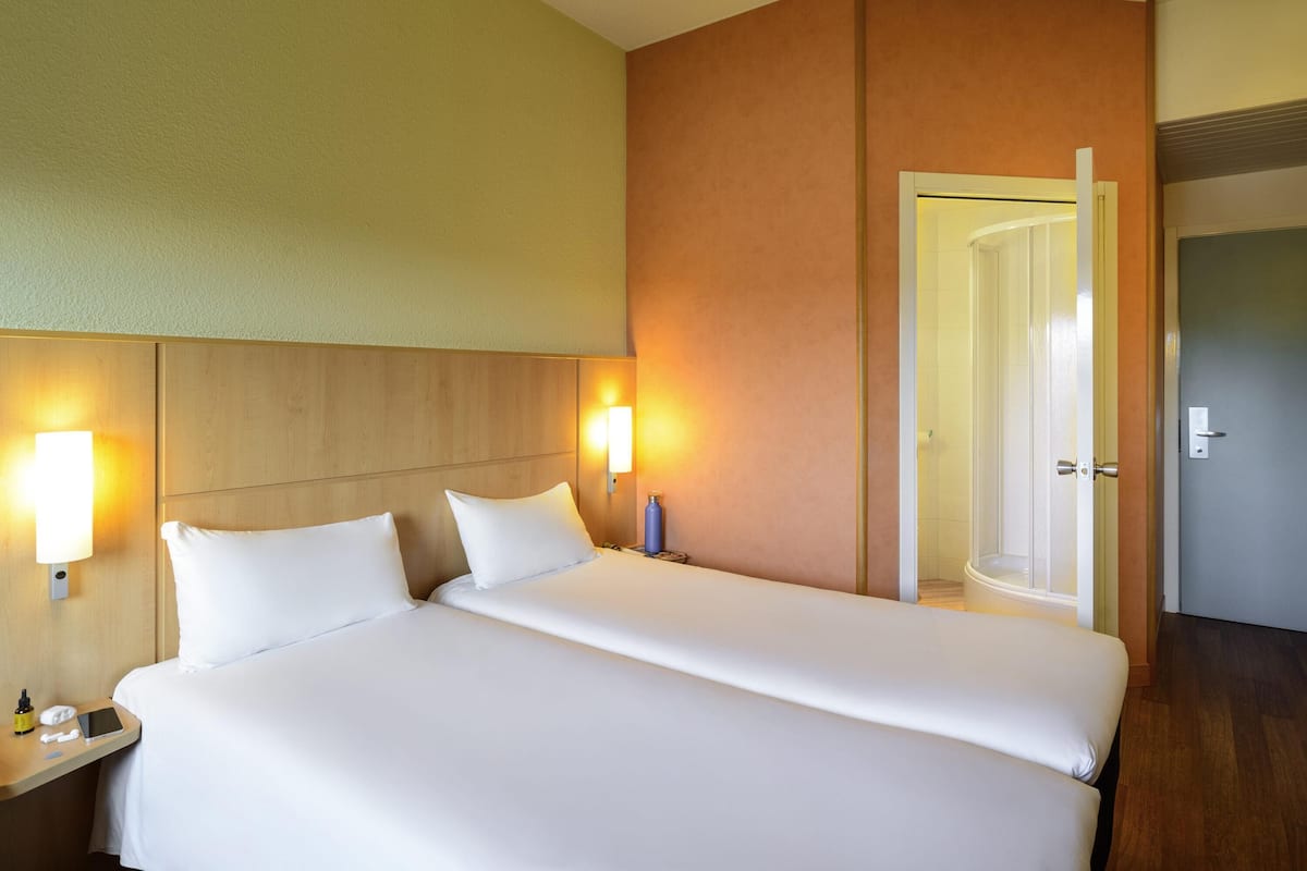 Una habitación de hotel con una cama grande, dos lámparas de noche, una mesita de noche con un teléfono y una botella, y una puerta que da a otra habitación.