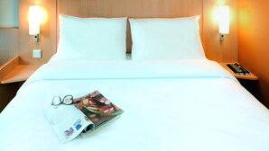 Een hotelkamer met een houten hoofdeinde, twee witte kussens, een netjes opgemaakt bed, een nachtkastje met een lamp, en een tijdschrift met brillen erop.