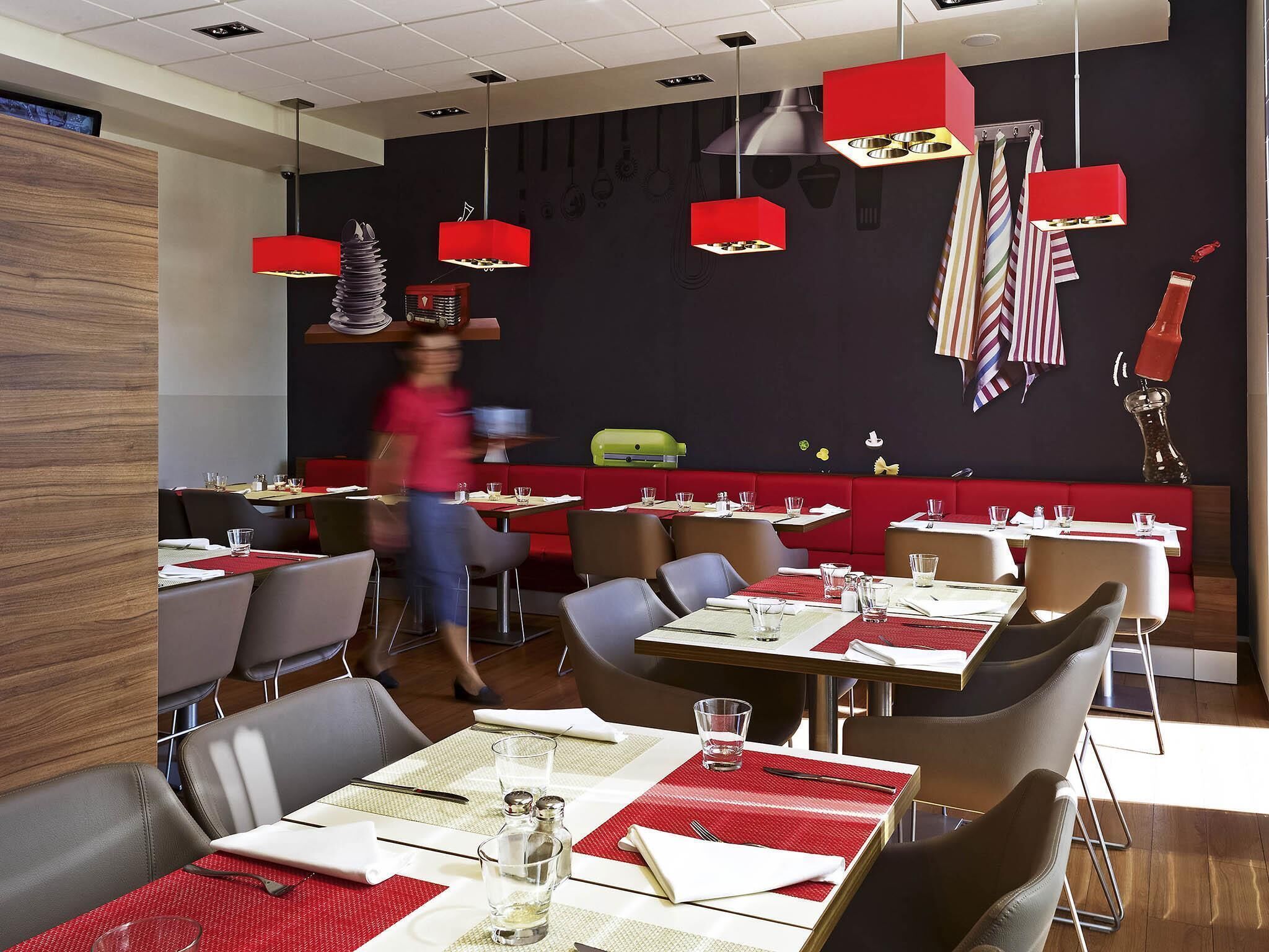 A modern restaurant interior with red accents, multiple dining tables, and a variety of hanging kitchen utensils on the wall.